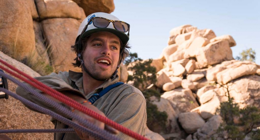 A person wearing a helmet braces themselves on taught ropes. There is a rock formation in the background. 